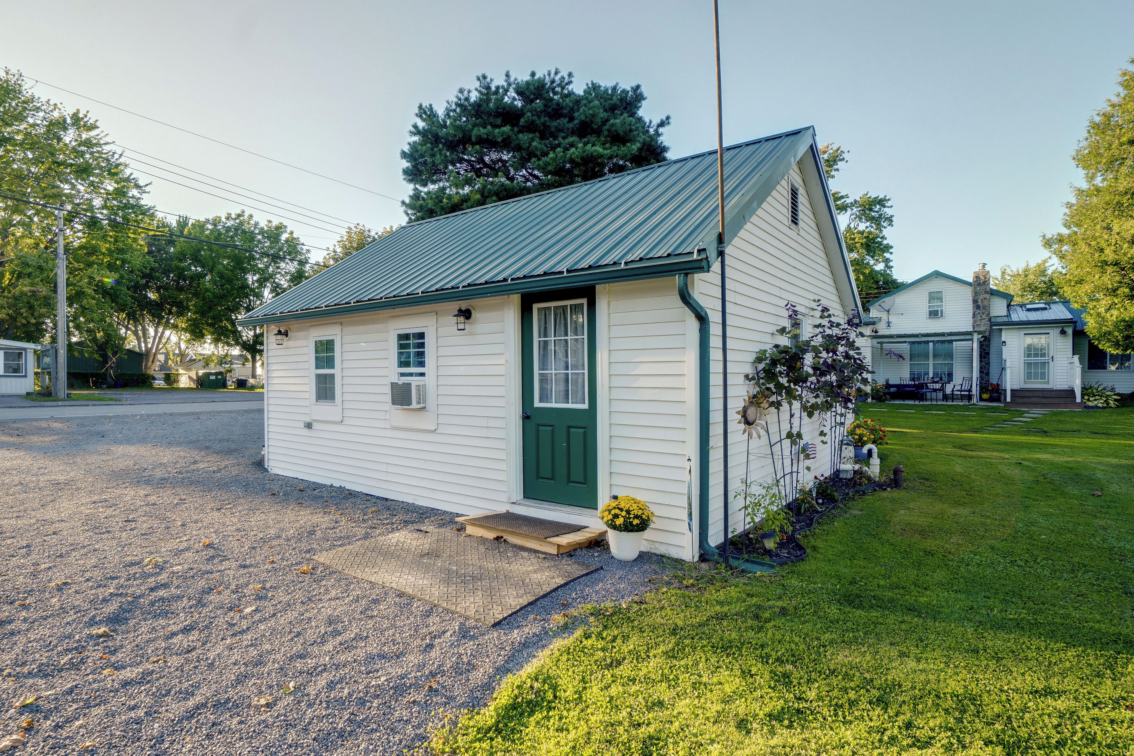 A small, single-story building with a green door and white siding, located on a grassy area with a gravel driveway.