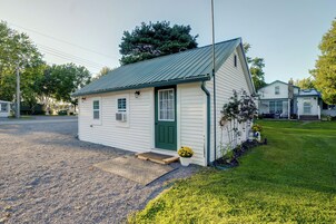 Um pequeno edifício térreo com porta verde e revestimento branco, situado em área gramada com entrada de cascalho.