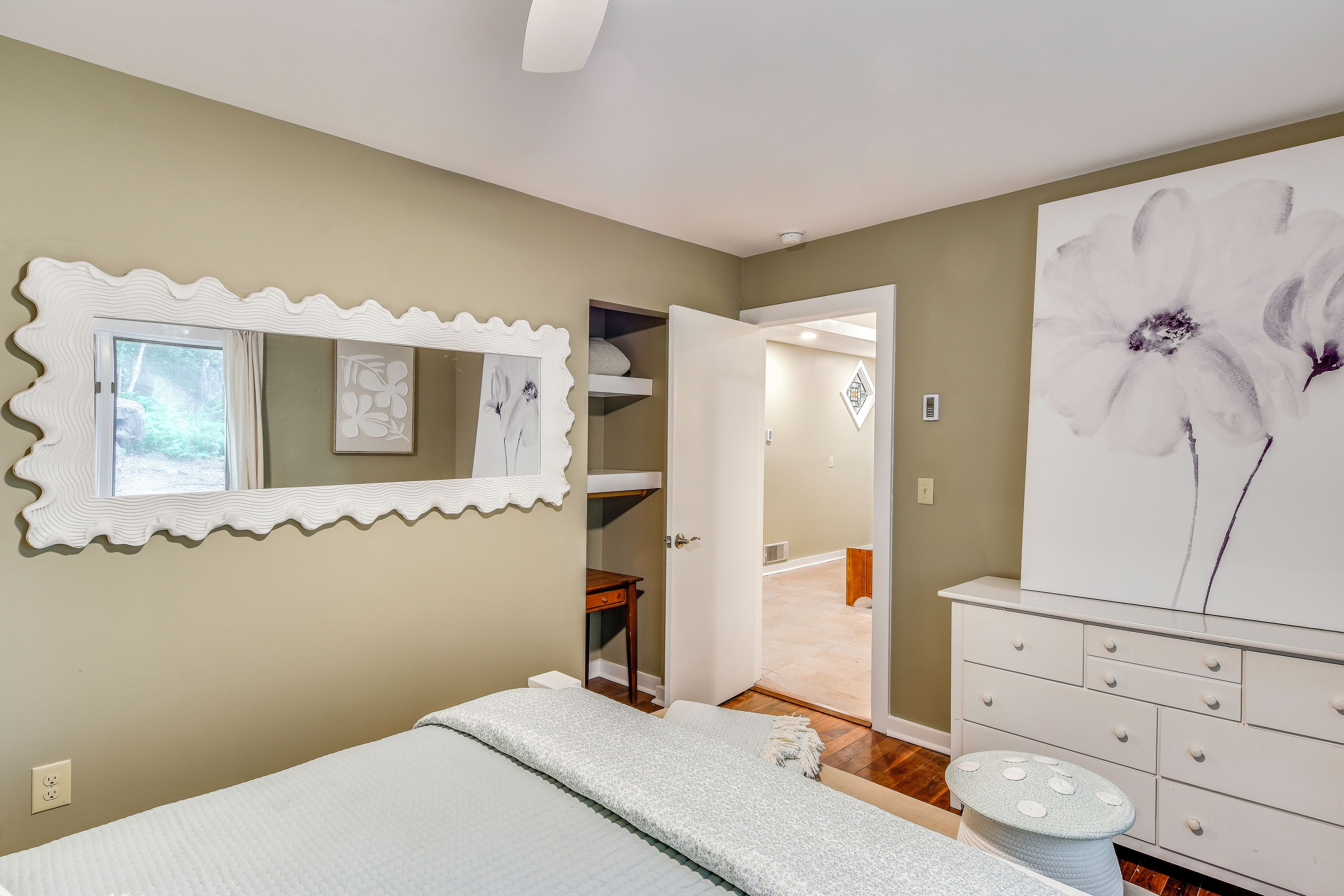 A bedroom with a bed, a dresser, a large floral painting, and a mirror with a wavy frame.
