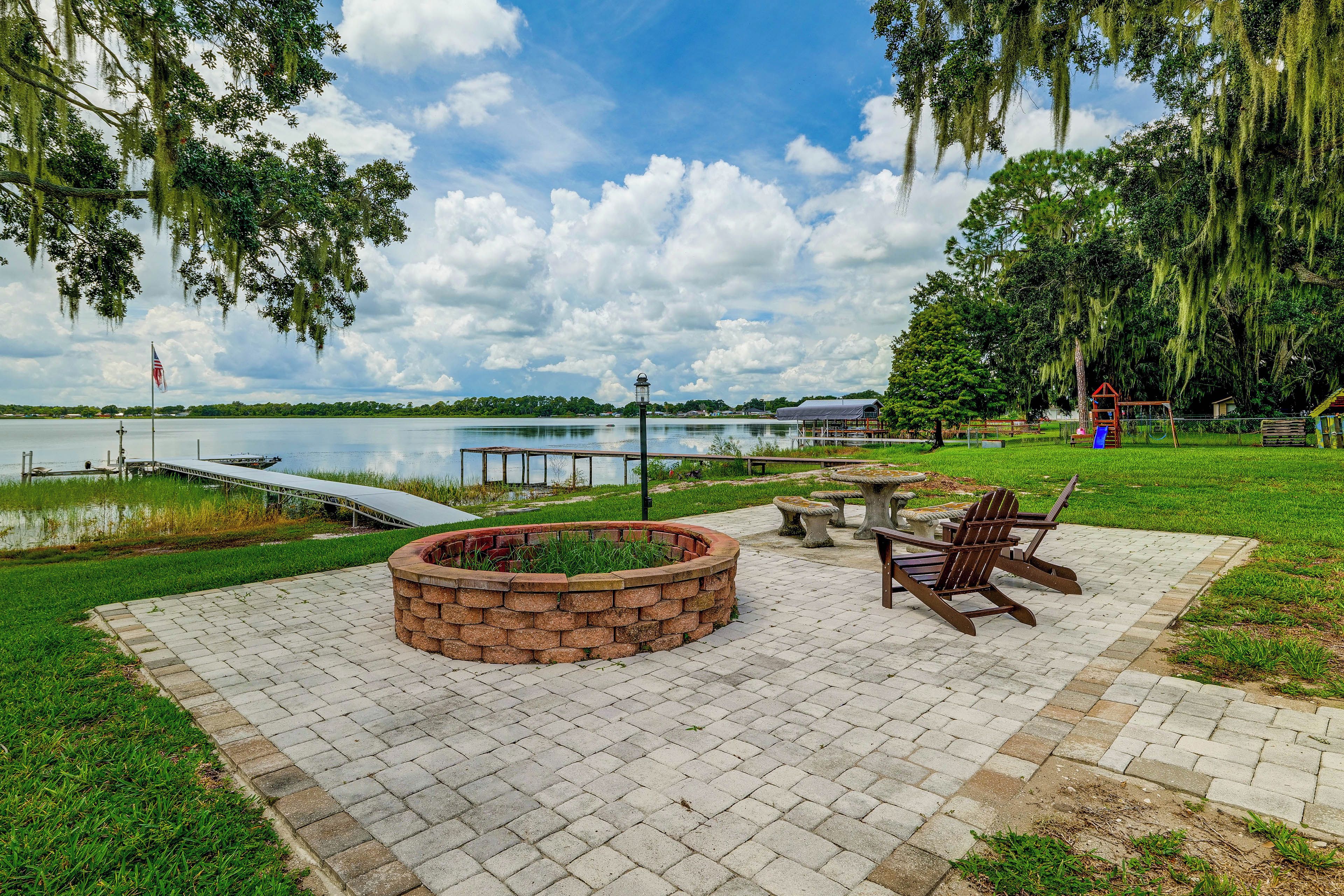 A lakeside park with a fire pit, picnic table, and Adirondack chairs.