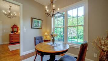 A dining room with a round table, chairs, a chandelier, a window with a view of greenery, and a framed picture on the wall.