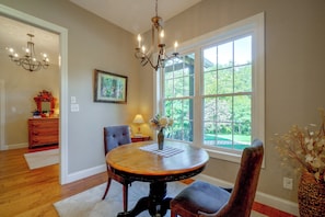 A dining room with a round table, chairs, a chandelier, a window with a view of greenery, and a framed picture on the wall.
