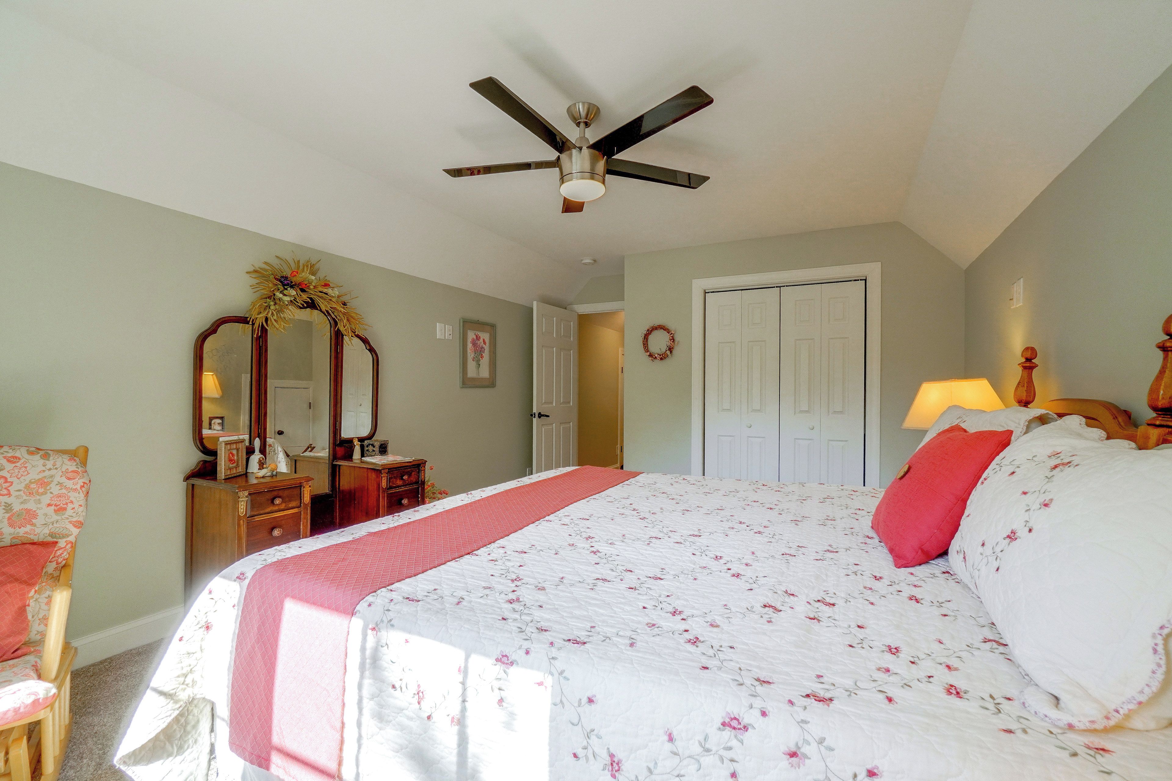 Une chambre à coucher comprenant un lit, une commode, un miroir, un ventilateur de plafond et une porte.