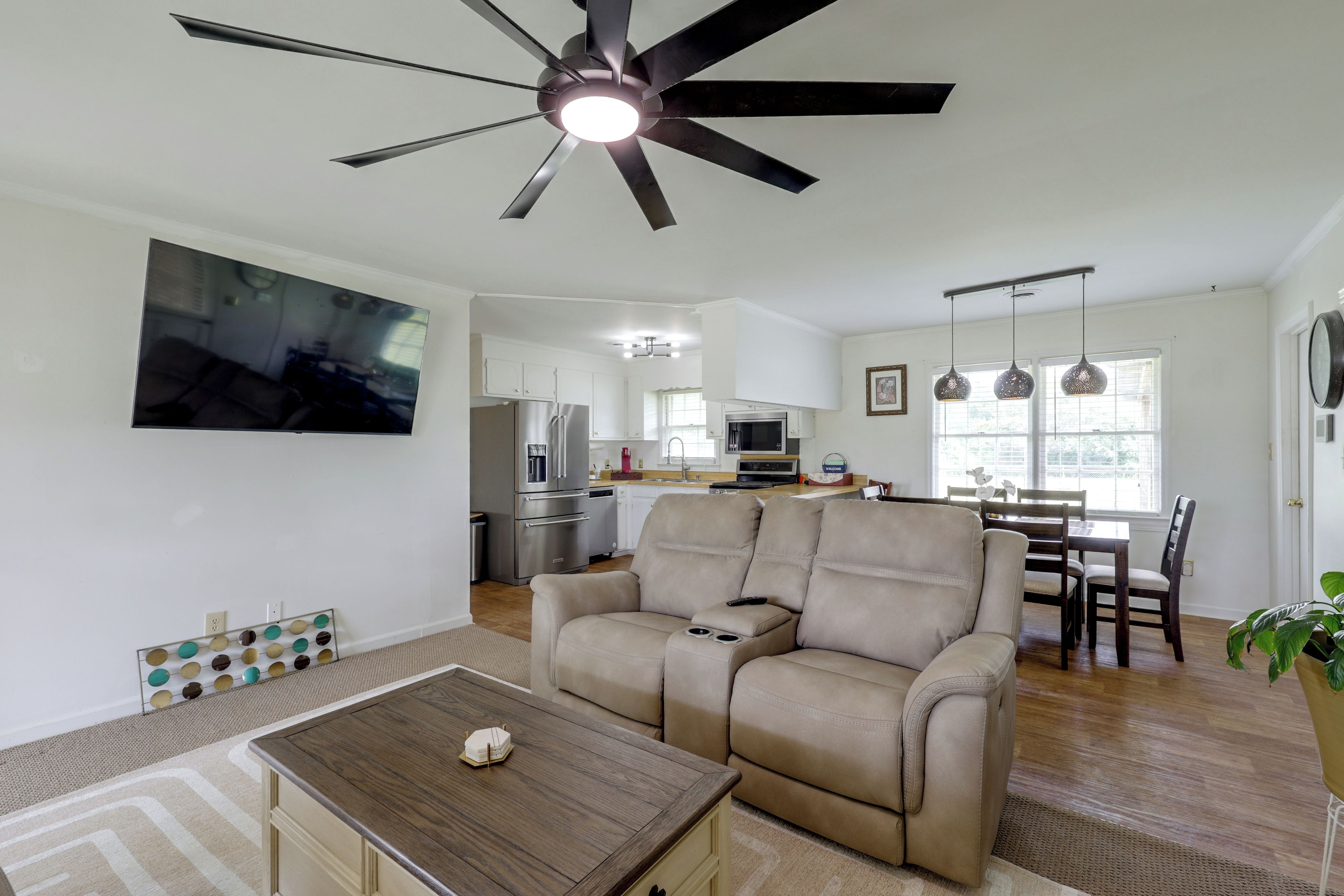 A modern living room with a ceiling fan, a flat-screen TV mounted on the wall, a beige sofa, a wooden coffee table, and a dining area with a table and chairs.