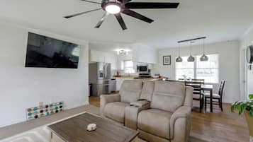 A modern living room with a ceiling fan, a flat-screen TV mounted on the wall, a beige sofa, a wooden coffee table, and a dining area with a table and chairs.