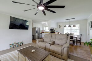 Una sala de estar moderna con ventilador de techo, un televisor de pantalla plana en la pared, un sofá color beige, una mesa de centro de madera y un comedor con mesa y sillas.