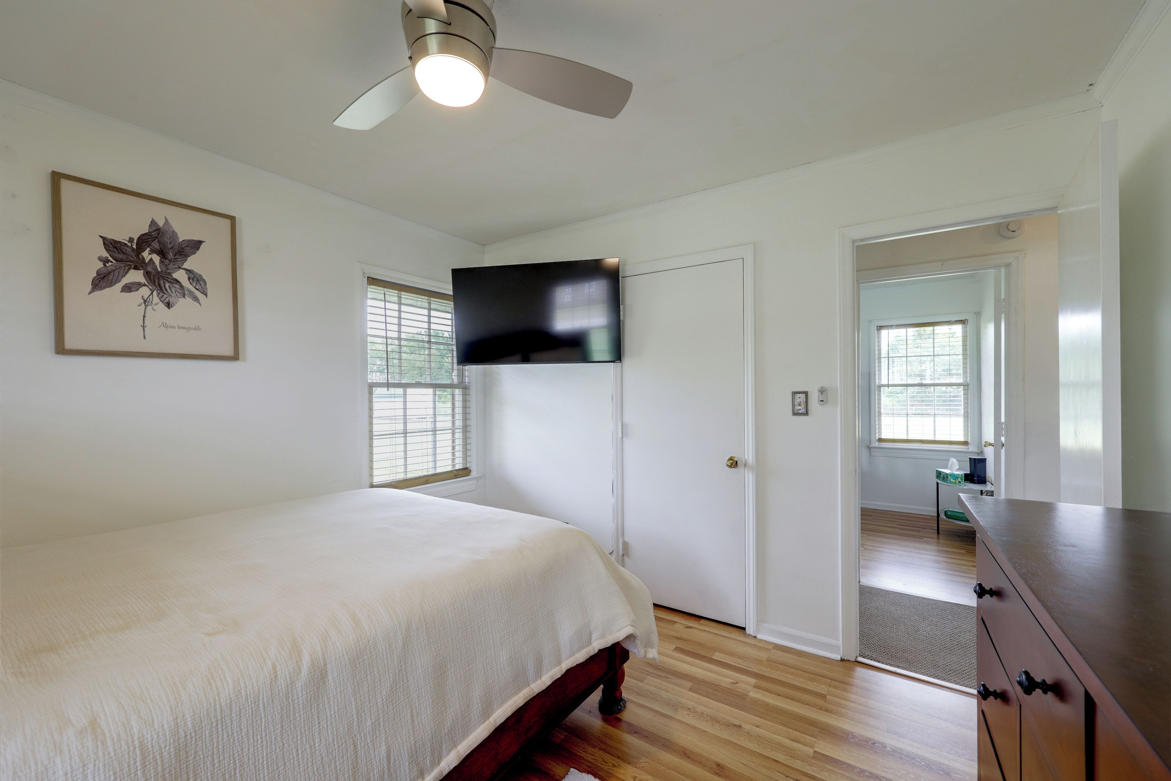Une chambre à coucher comprenant un lit, un ventilateur de plafond, un téléviseur fixé au mur et une porte donnant sur une autre pièce.