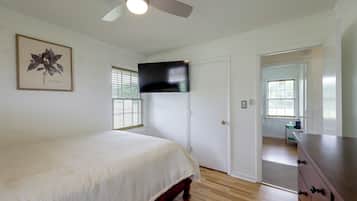 Une chambre à coucher comprenant un lit, un ventilateur de plafond, un téléviseur fixé au mur et une porte donnant sur une autre pièce.