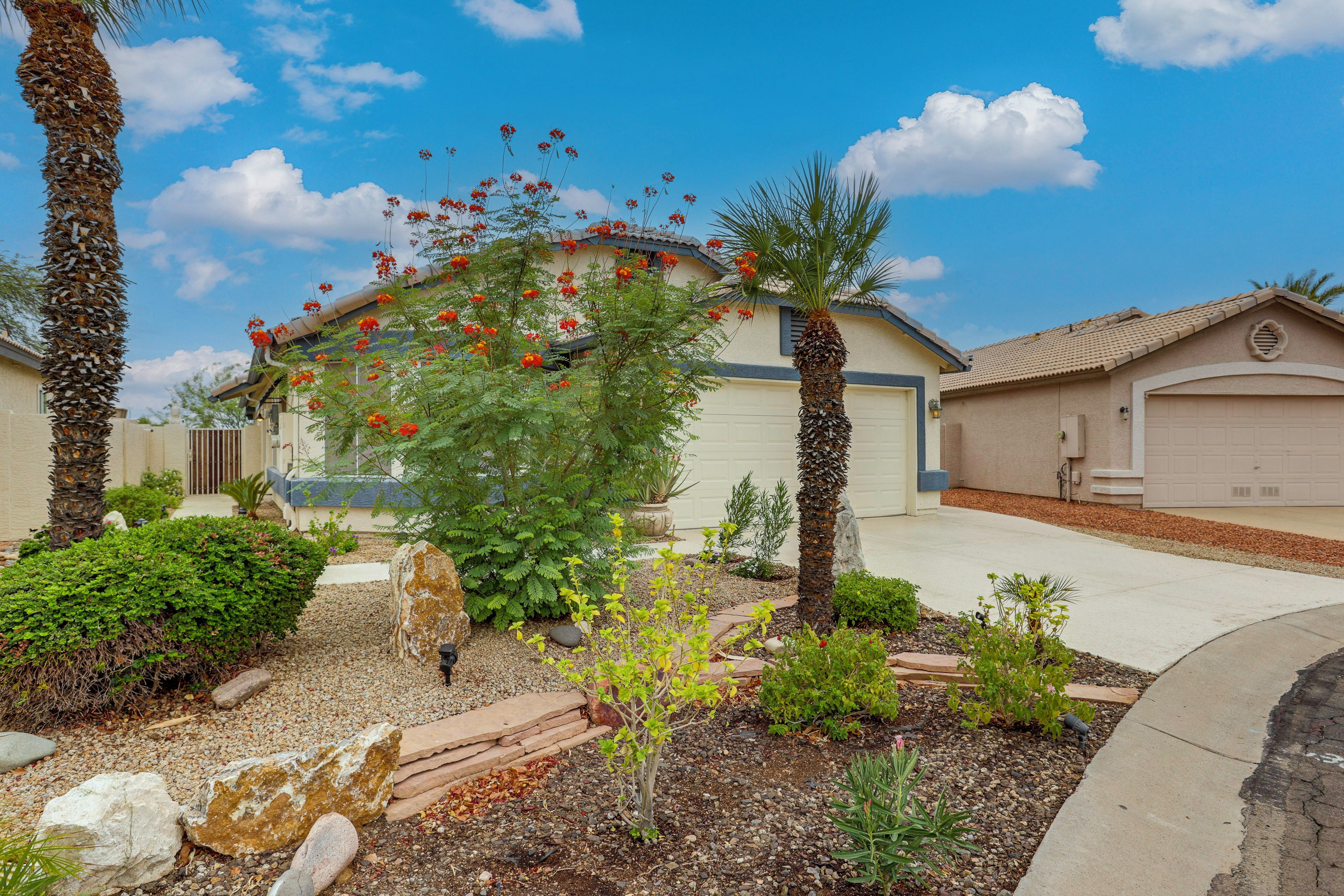 A residential area with a house featuring a garage, a driveway, and a well-maintained garden with various plants and a palm tree.