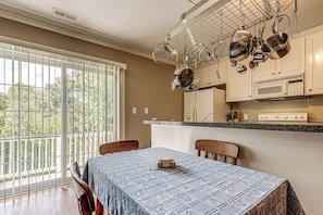 A dining area with a table set for two, a kitchen with hanging pots, and a view through a large window. - Raleigh-area Farm Stay w/ Pond: Swim, Fish, Relax! (Clayton)
