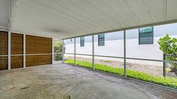 Un espacio de patio cubierto con suelo de piedra, listones de madera y ventanas amplias que dan vistas a zonas verdes y un edificio.