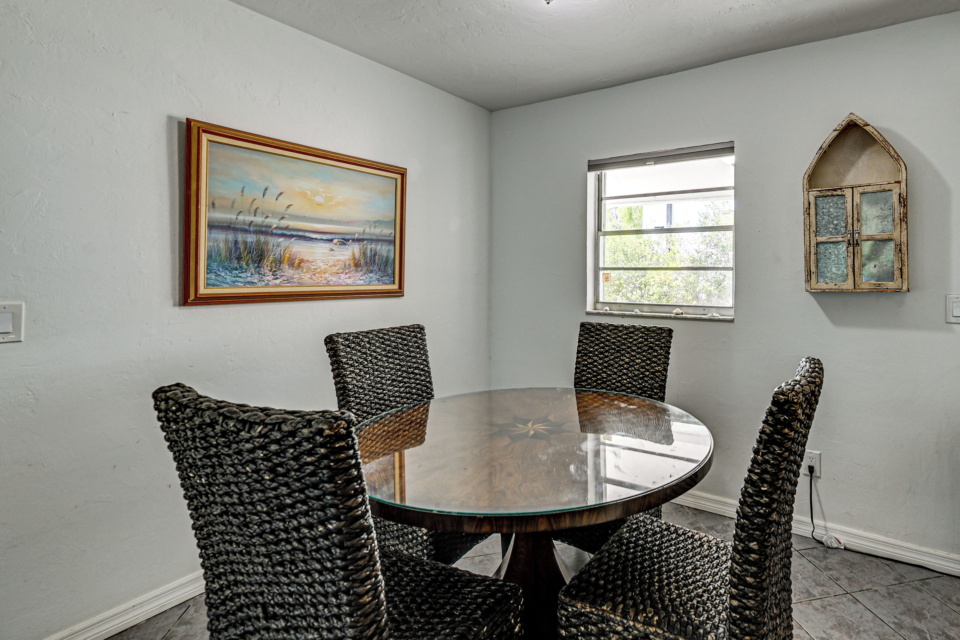 A dining area with a round table and woven chairs, a painting on the wall, a window with a view, and a decorative arched mirror.