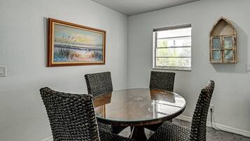 A dining area with a round table and woven chairs, a painting on the wall, a window with a view, and a decorative arched mirror.