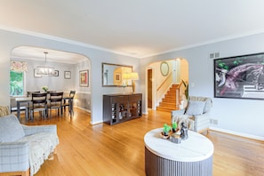 A spacious living room with a wooden floor, a dining area with a table and chairs, a staircase, and a large painting on the wall.