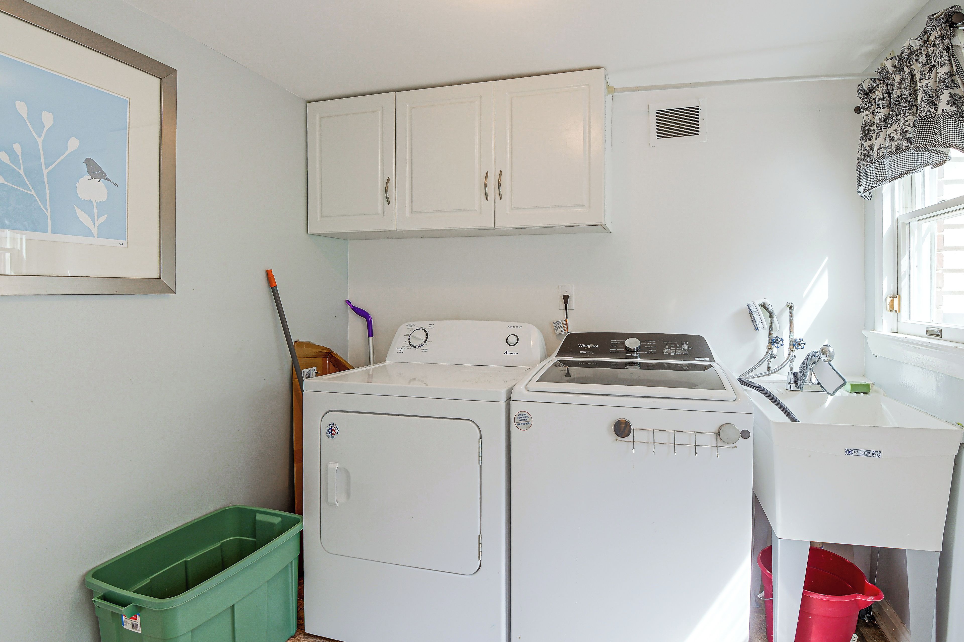 A laundry room with a washer and dryer, a green trash bin, and a window with curtains.