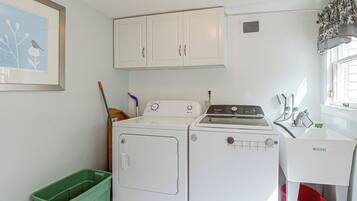 A laundry room with a washer and dryer, a green trash bin, and a window with curtains.