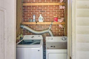 Uma lavanderia com duas máquinas de lavar, uma prateleira com produtos de limpeza e uma mangueira de ventilação.