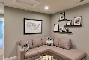 Una sala de estar con un sofá seccional color beige, un mapa enmarcado de los Estados Unidos y varias fotografías enmarcadas en una pared.