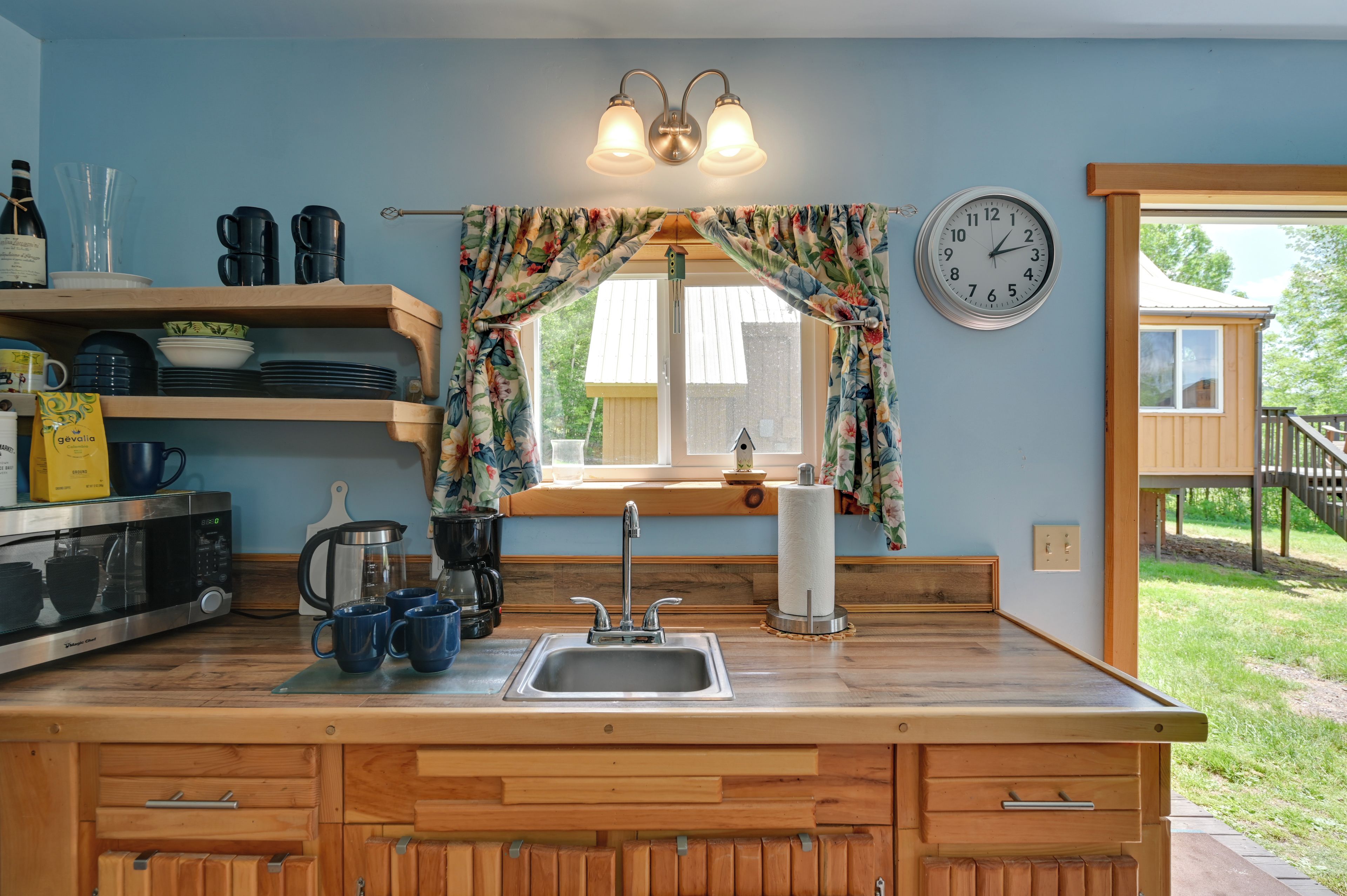 A kitchen with wooden cabinets, a sink, and a window with curtains. There is a clock on the wall and a wooden deck outside.