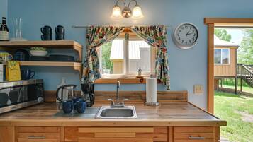 A kitchen with wooden cabinets, a sink, and a window with curtains. There is a clock on the wall and a wooden deck outside.