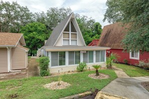 Une maison d’habitation avec un toit à pignon, un porche à l’avant et un petit jardin devant, agrémenté d’un palmier et de quelques arbustes.
