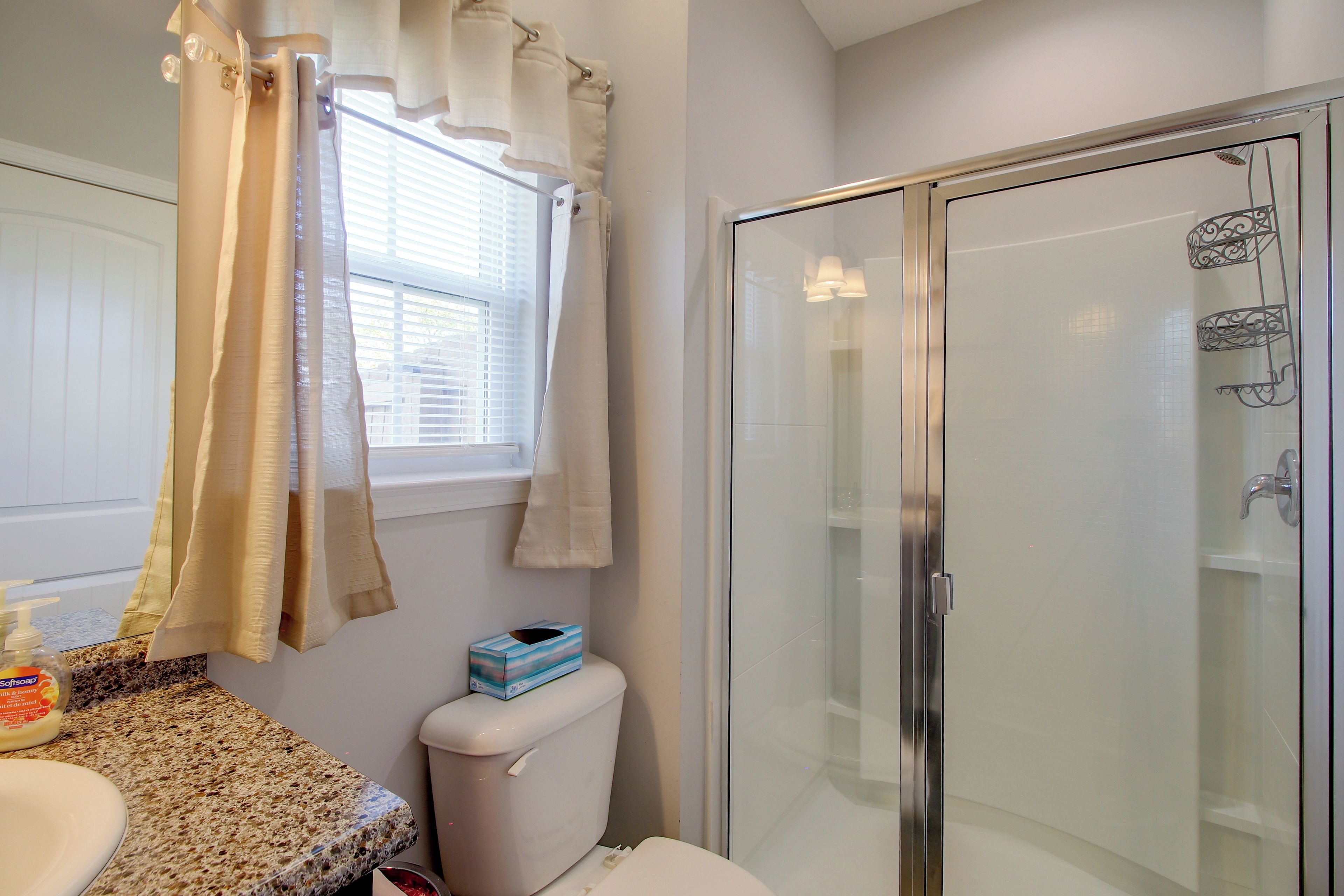 Une salle de bain comprenant des toilettes, un espace de douche vitré, une fenêtre avec stores et un plan de toilette doté d’un distributeur de savon.