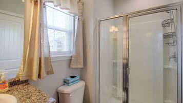 Une salle de bain comprenant des toilettes, un espace de douche vitré, une fenêtre avec stores et un plan de toilette doté d’un distributeur de savon.