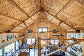 Interior de una cabaña con techo alto, vigas de madera, zona de comedor con mesa y sillas, un sillón colgante y una pared de piedra.