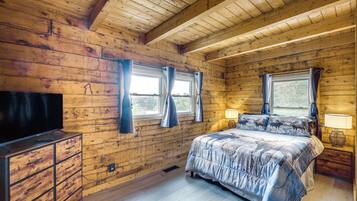 A wooden cabin bedroom with a bed, two windows with curtains, a TV, and a bedside lamp.
