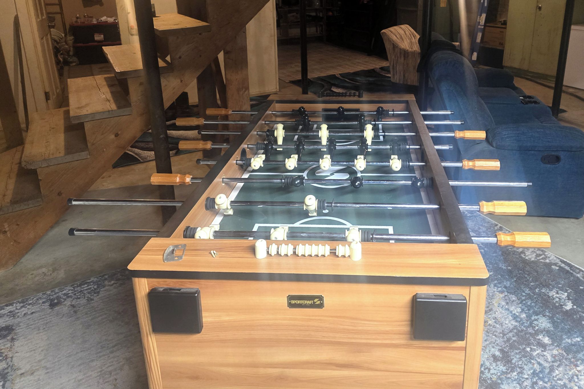 A foosball table with a wooden base and black accents, located in a room with a concrete floor and wooden furniture.
