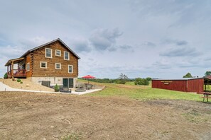 Una cabaña de madera con un porche, un patio con mobiliario exterior y un granero rojo al fondo.