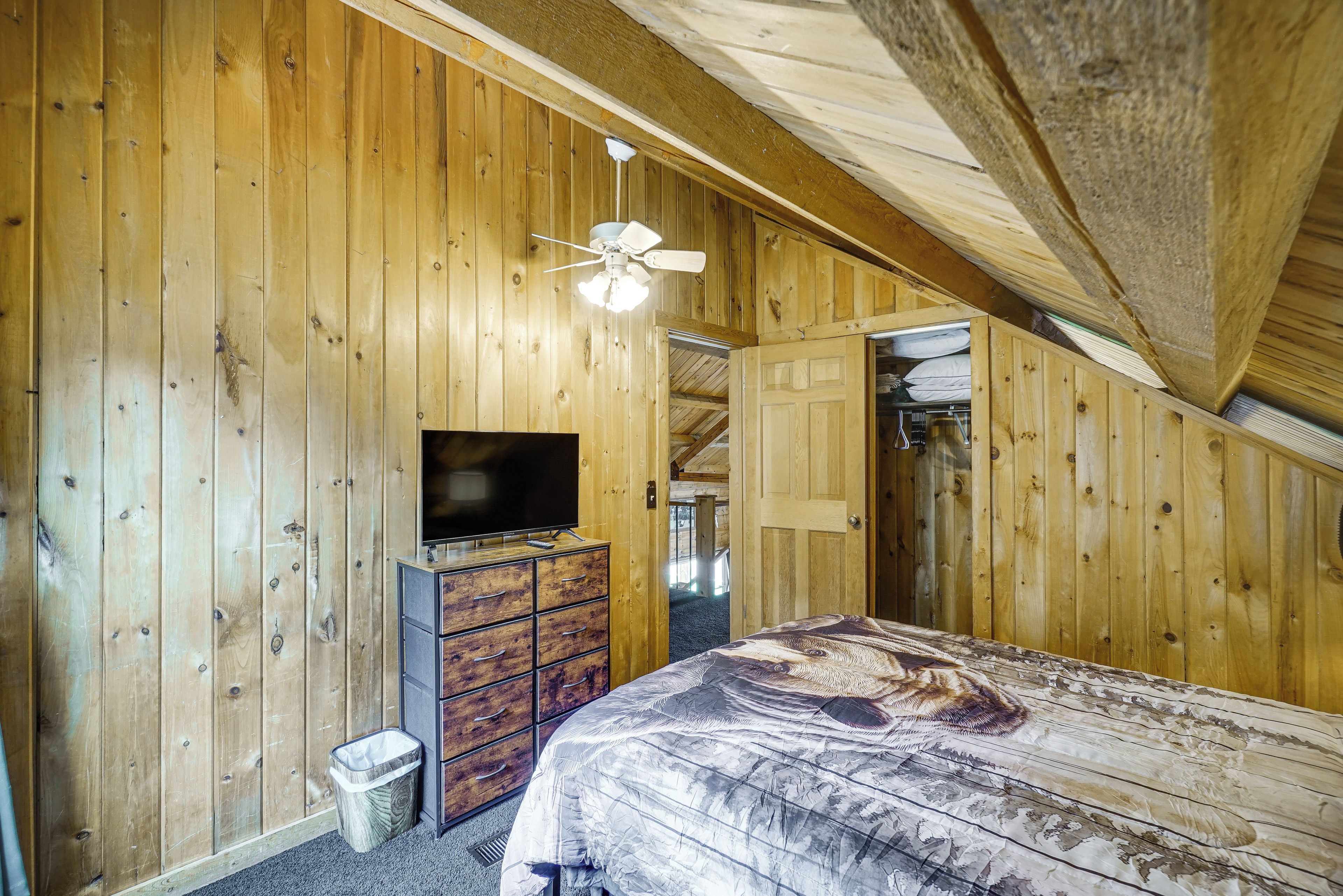 A room with wooden walls, a ceiling fan, a flat-screen TV, a bed with a patterned bedspread, and a dresser.