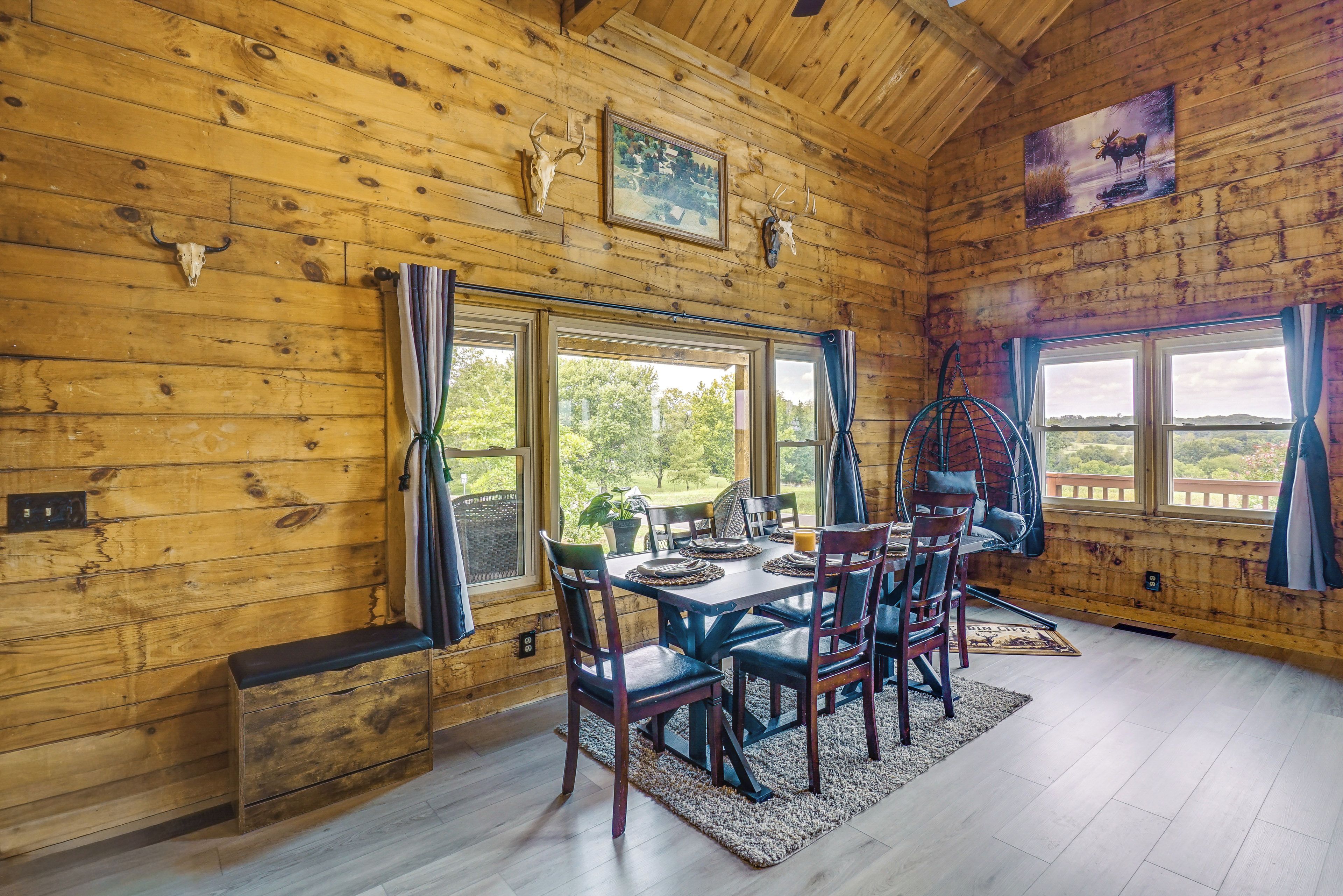 A wooden cabin interior with a dining table set for four, a bench, and a view of the outdoors through large windows.