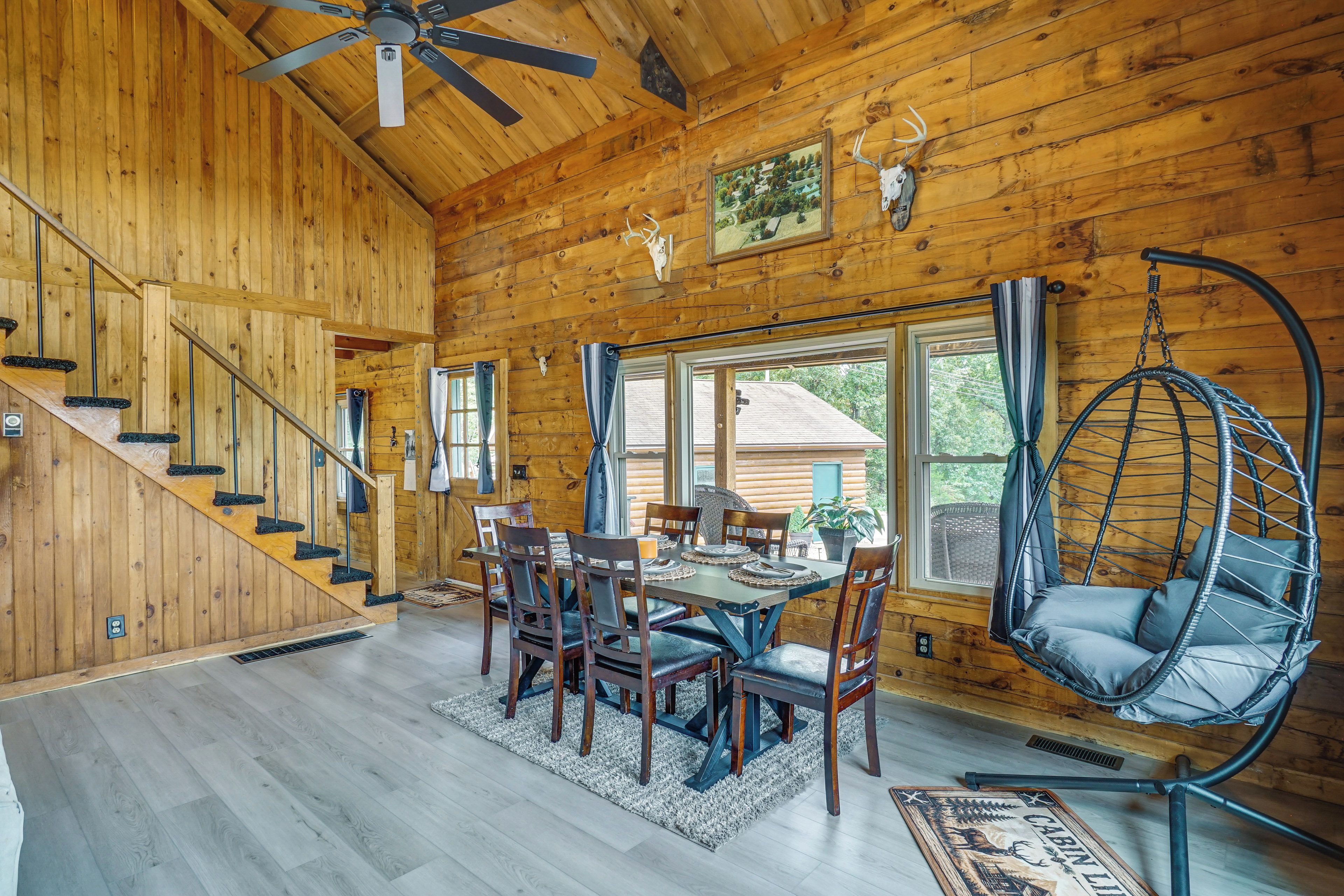 A wooden cabin interior with a dining area, a hanging chair, and a staircase.