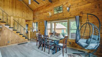 A wooden cabin interior with a dining area, a hanging chair, and a staircase.