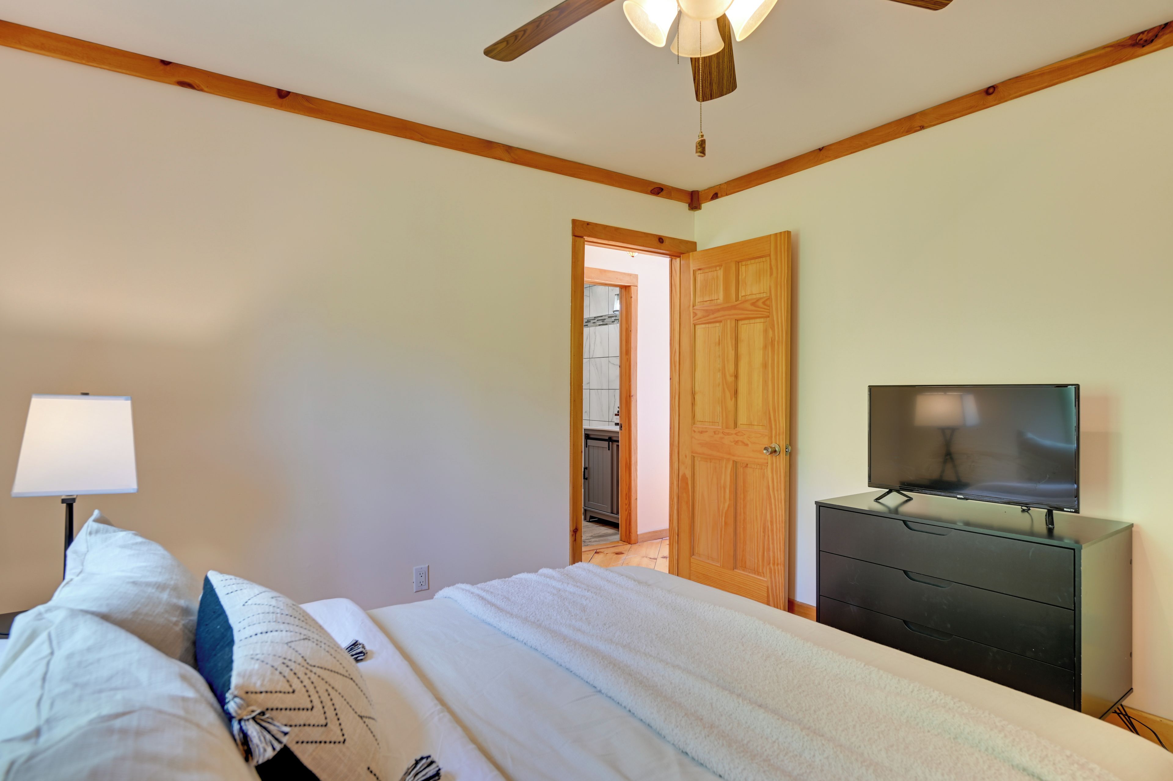 A bedroom with a bed, a TV on a dresser, a ceiling fan, and a wooden door leading to a kitchen.