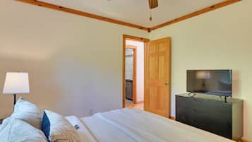 A bedroom with a bed, a TV on a dresser, a ceiling fan, and a wooden door leading to a kitchen.