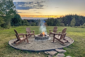 A circular fire pit with four Adirondack chairs around it, set in a grassy area with a view of a forested landscape and a sunset in the background.