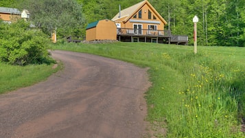 A wooden cabin with a deck, surrounded by lush greenery and a dirt road.