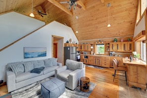 A spacious living area with a gray sofa, wooden ceiling beams, and a kitchen with wooden cabinets.