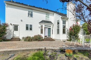 A two-story white house with a brick driveway, a front porch with steps, and a small garden area with a bird bath and chair.