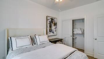 A bedroom with a bed, a desk, a chair, a ceiling fan, a wardrobe, and a painting on the wall.