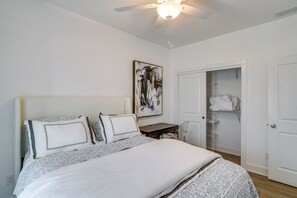 A bedroom with a bed, a desk, a chair, a ceiling fan, a wardrobe, and a painting on the wall.