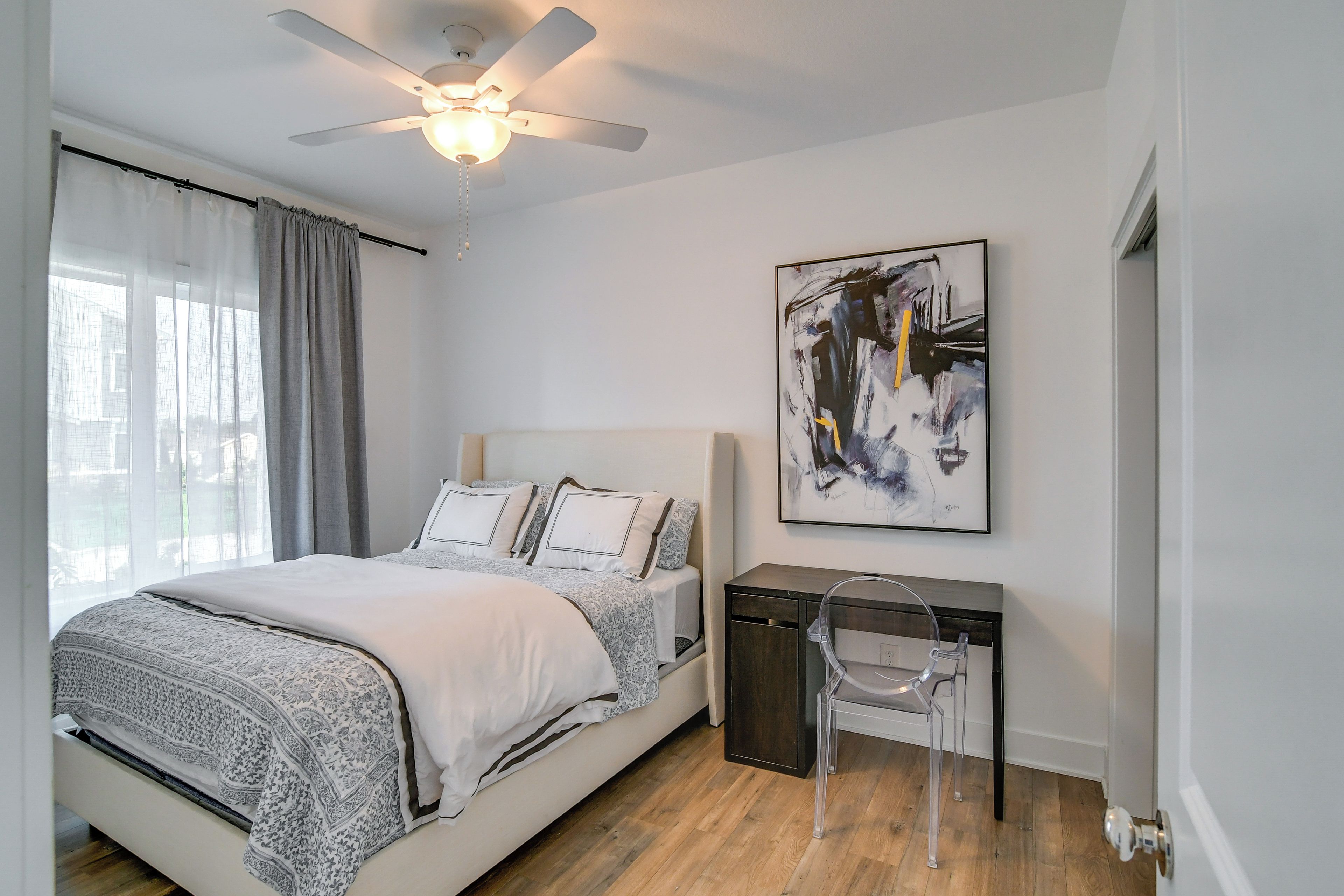 A bedroom with a bed, a desk, a chair, a ceiling fan, and a painting on the wall.
