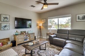 A living room with a flat-screen TV, a leather sofa, a coffee table, a bookshelf, and a window with a view of palm trees. - Private Dock & Pool: Waterfront Key Largo Home (Key Largo)