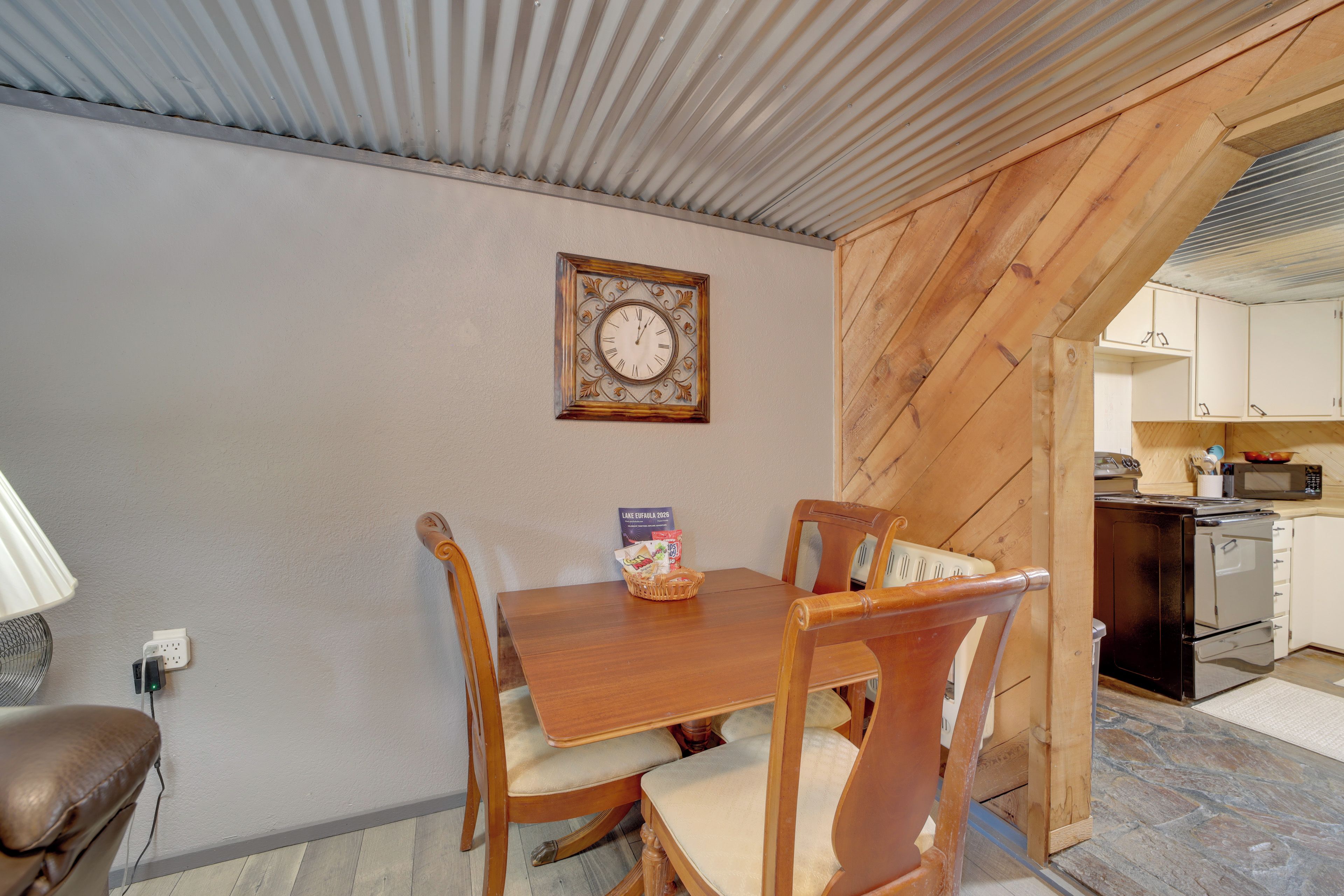 Un comedor con mesa y sillas de madera, un reloj de pared y una cocina visible a través de un umbral abierto.