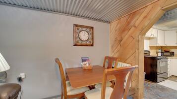 Un comedor con mesa y sillas de madera, un reloj de pared y una cocina visible a través de un umbral abierto.