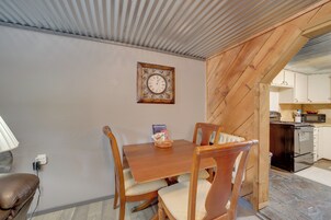 Un comedor con mesa y sillas de madera, un reloj de pared y una cocina visible a través de un umbral abierto.