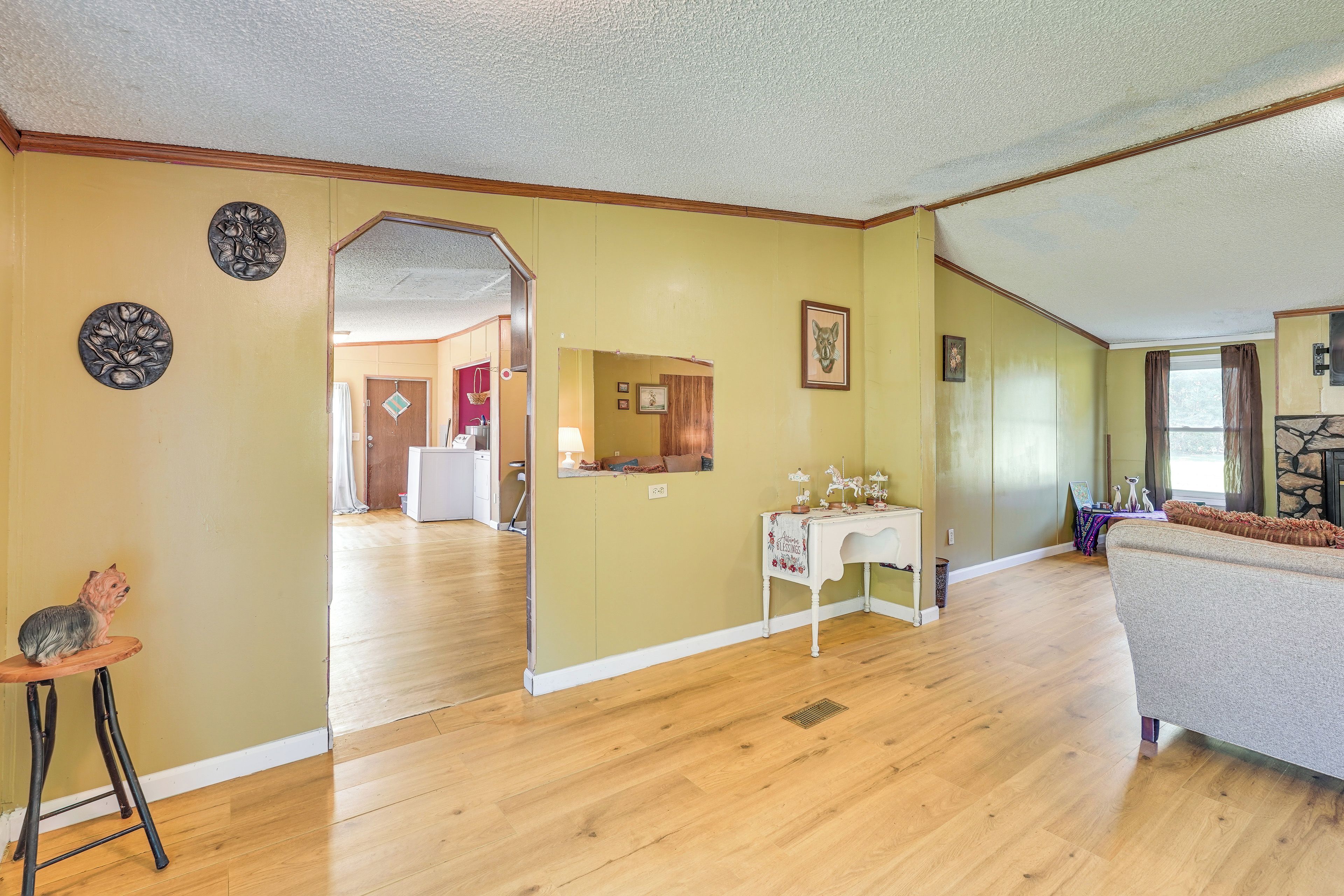 Uma sala de estar ampla com piso de madeira, um sofá branco, uma mesa pequena com objetos decorativos e uma área de cozinha visível através de uma porta aberta.