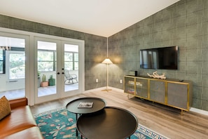 Uma sala de estar moderna com um sofá, duas mesas pretas, um abajur, uma televisão e uma parede com padrão.